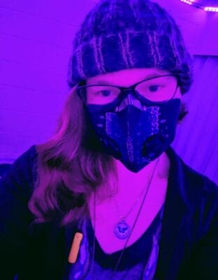 Self-portrait of a person wearing a patterned face mask, black beanie, and glasses under purple neon lighting, with a pendant necklace visible.