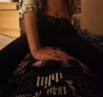 Person sitting on a bed, midriff showing, with a belt and patterned shirt; a hand with rings rests on a plaid fabric in the foreground.