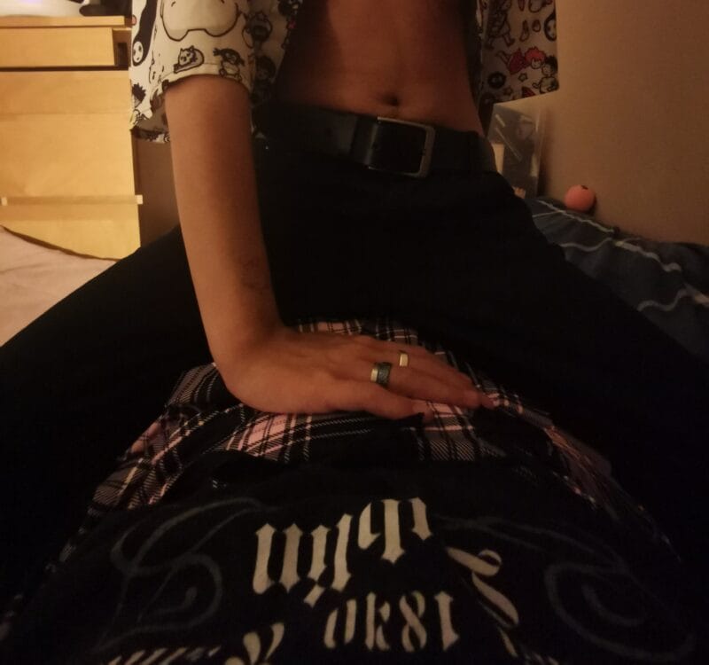 Person sitting on a bed, midriff showing, with a belt and patterned shirt; a hand with rings rests on a plaid fabric in the foreground.
