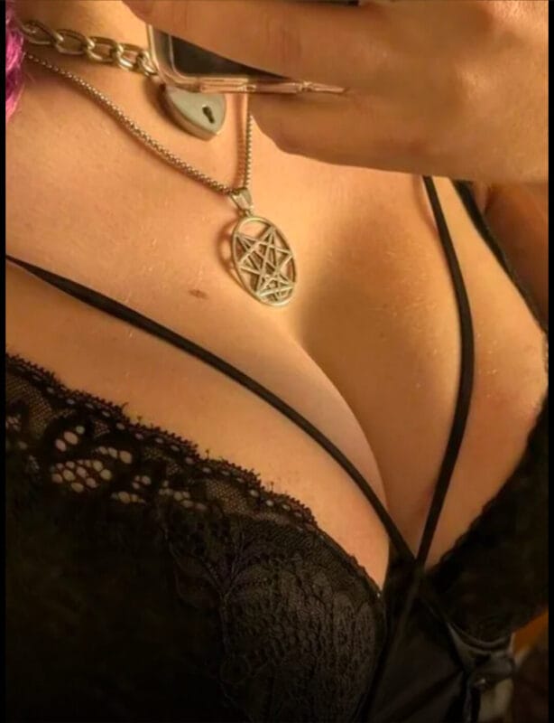 Close-up of a woman's chest wearing black lace lingerie and a silver pentagram pendant necklace, with a phone held near her face in the background.