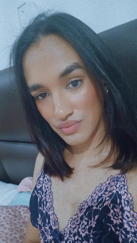 Close-up selfie of a woman with shoulder-length dark hair wearing a navy lace top, posing on a bed with a padded headboard in the background.