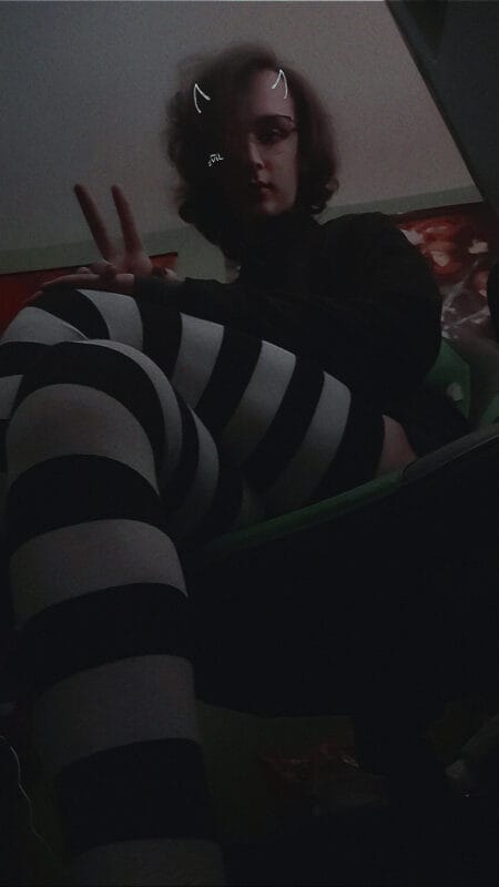 Person sitting in a dim room, wearing a black top and bold black-and-white striped tights, making a peace sign toward the camera.