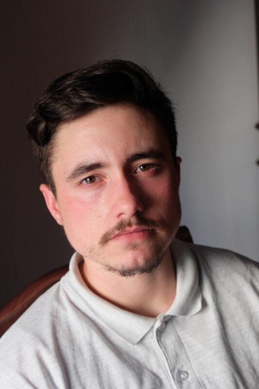 Portrait of a young man with dark hair and light stubble looking at the camera indoors, wearing a light gray polo shirt.